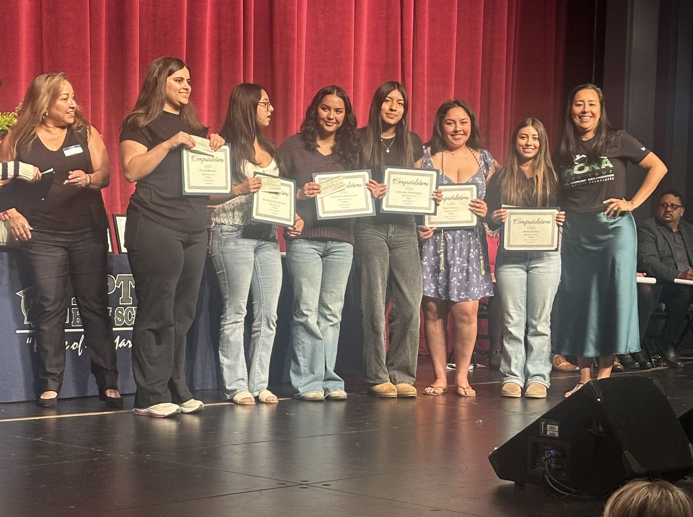 Scholarship recipients holding their certificates on stage at Aptos High School with NHORA Santa Cruz members